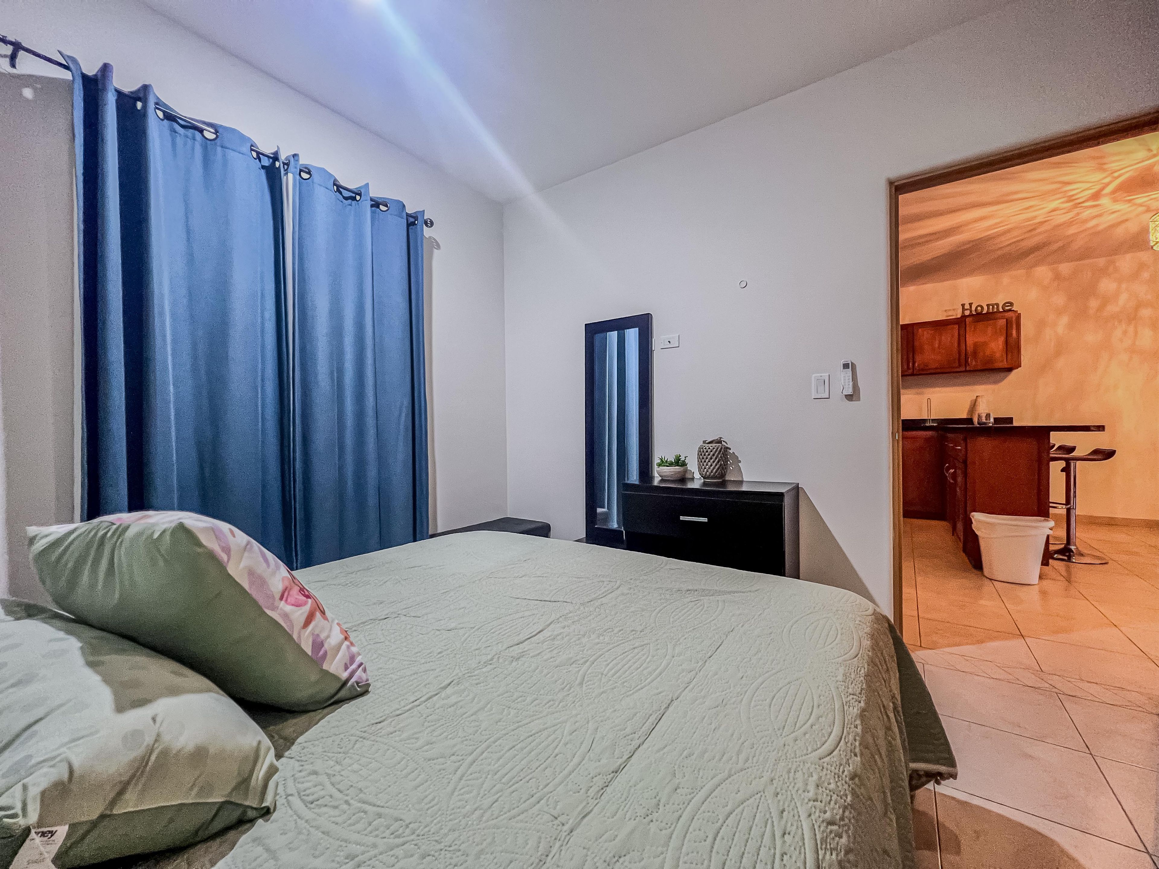 A bedroom with a bed, pillows, a nightstand, a mirror, and a view into a kitchen area.