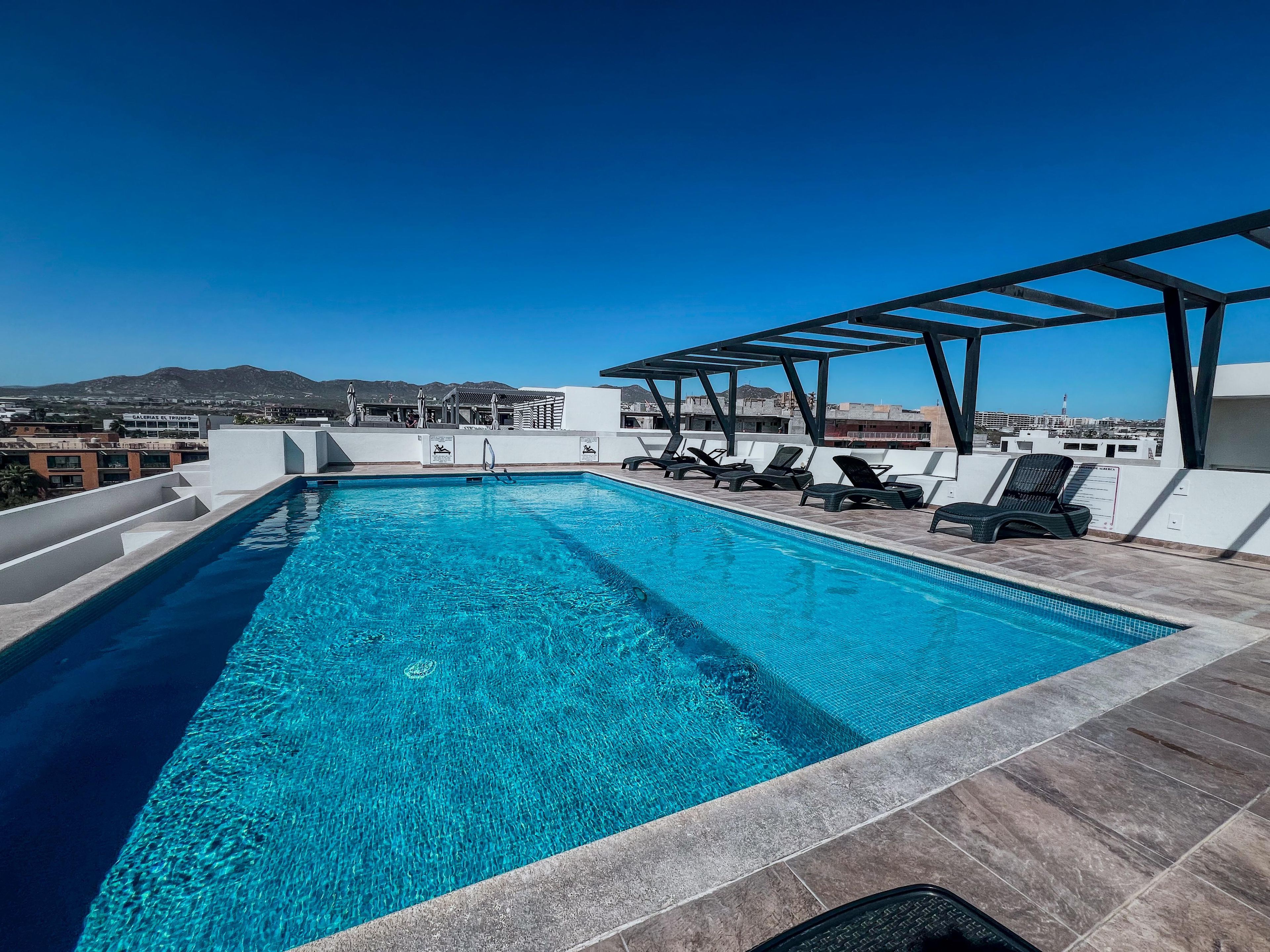 A rooftop pool with clear blue water, lounge chairs, and a pergola structure.