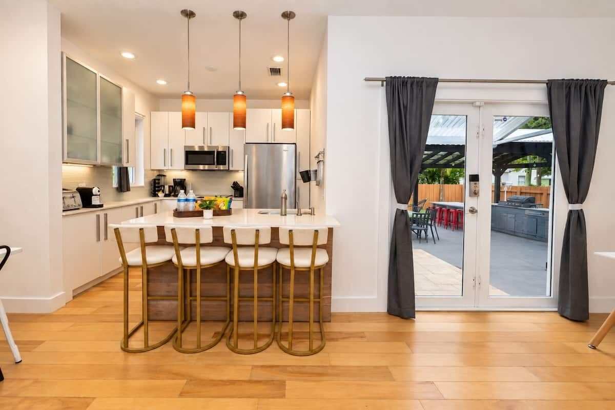 Een moderne keuken met een centraal eiland, barkrukken, hanglampen en een glazen schuifdeur die leidt naar een buitenruimte.