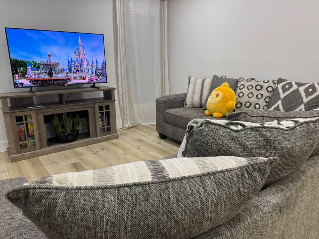 Una sala de estar con una televisión de pantalla plana, un sofá gris y un peluche amarillo.