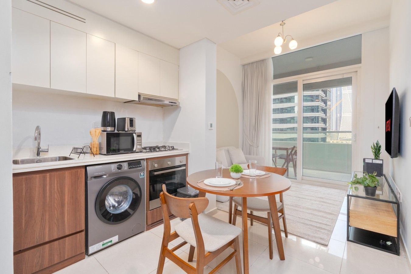 A compact kitchen with a washing machine, microwave, and stove. There is a dining area with a table and chairs, and a balcony with a view of the city.