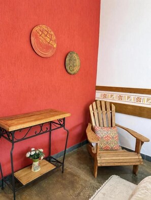 Een hoek met een rode muur, een houten stoel met een gekleurd kussen, een klein houten tafeltje met een bloevas, en twee decoratieve borden.