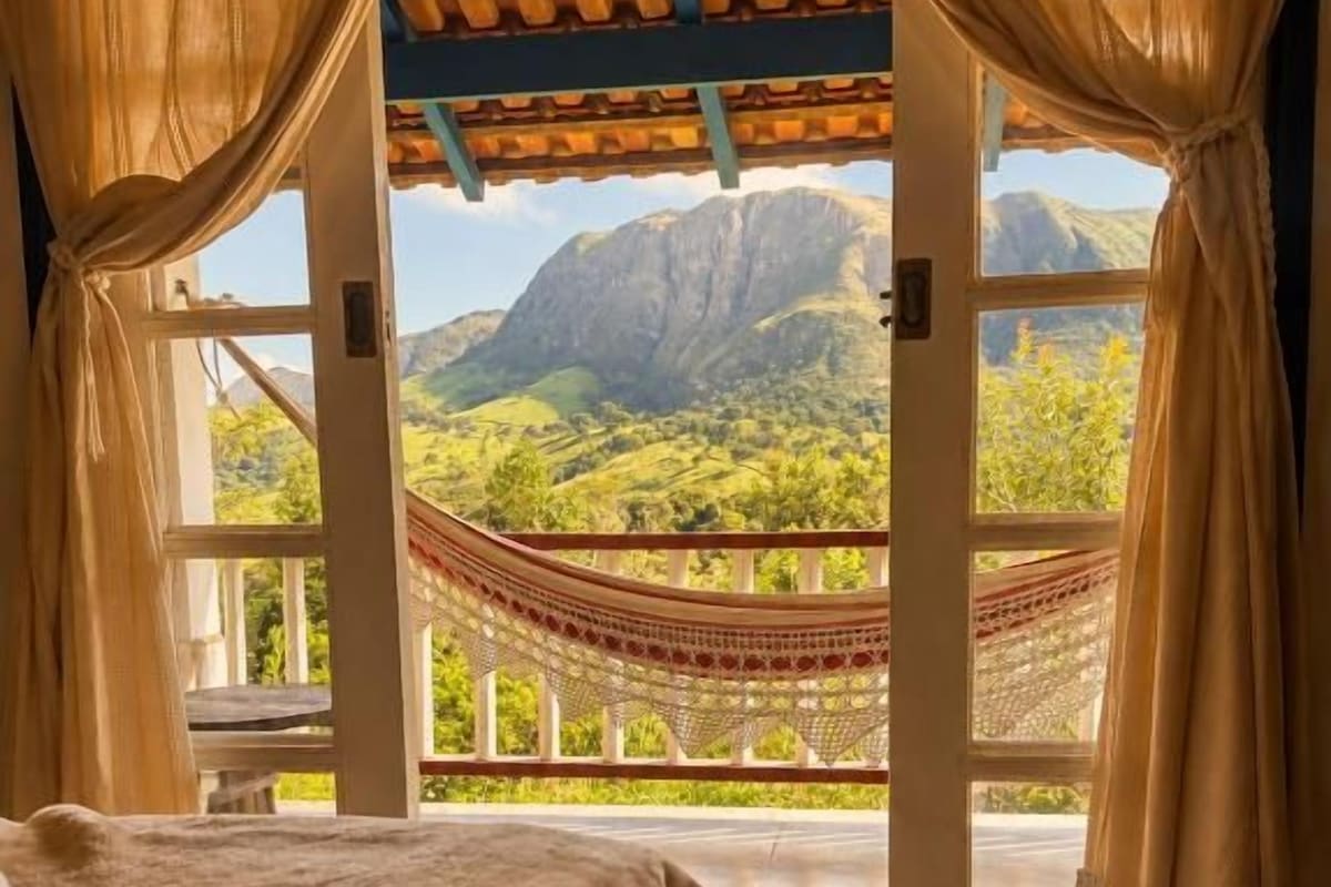 Una habitación con cama, una hamaca y vistas a montañas a través de una puerta abierta.