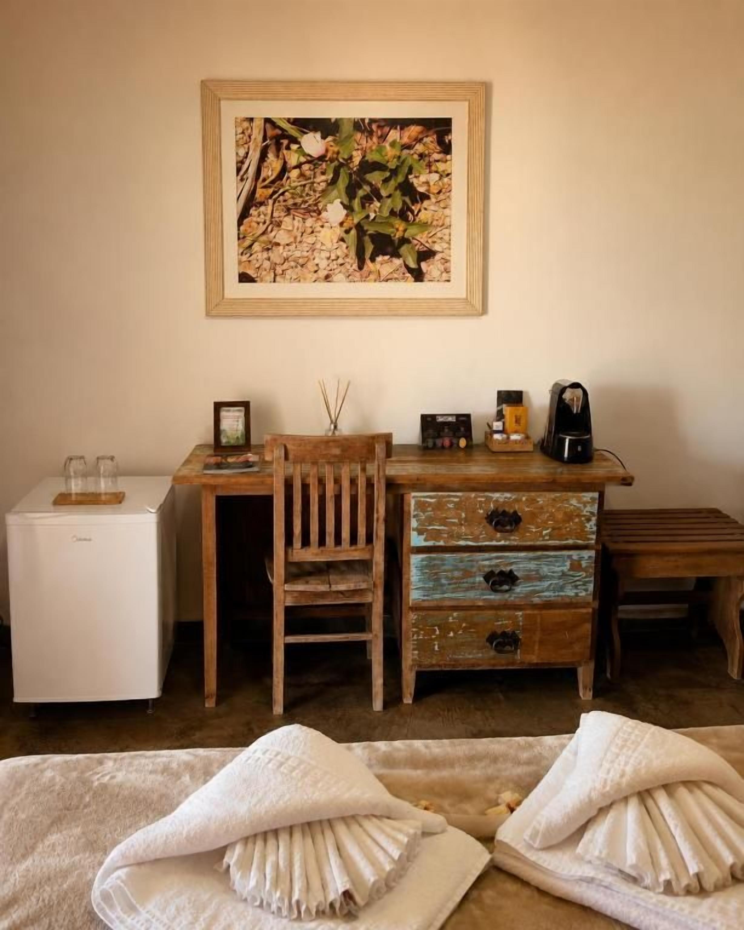 Una habitación con escritorio de madera, una silla, refrigerador pequeño, cafetera y un cuadro en la pared.