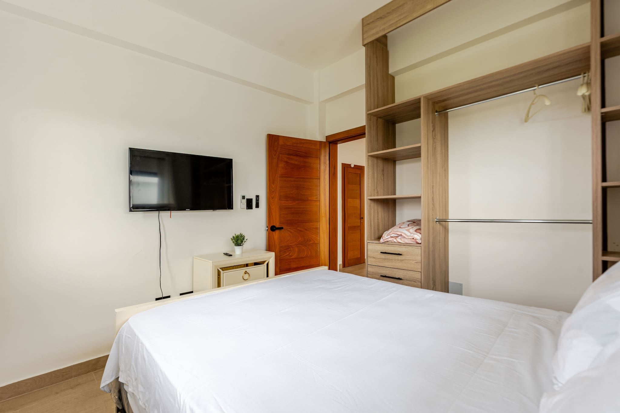 A bedroom with a bed, a TV mounted on the wall, a bedside table, a wooden wardrobe, and a mirror.