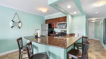 A kitchen with granite countertops, wooden chairs, a stainless steel refrigerator, and a microwave.