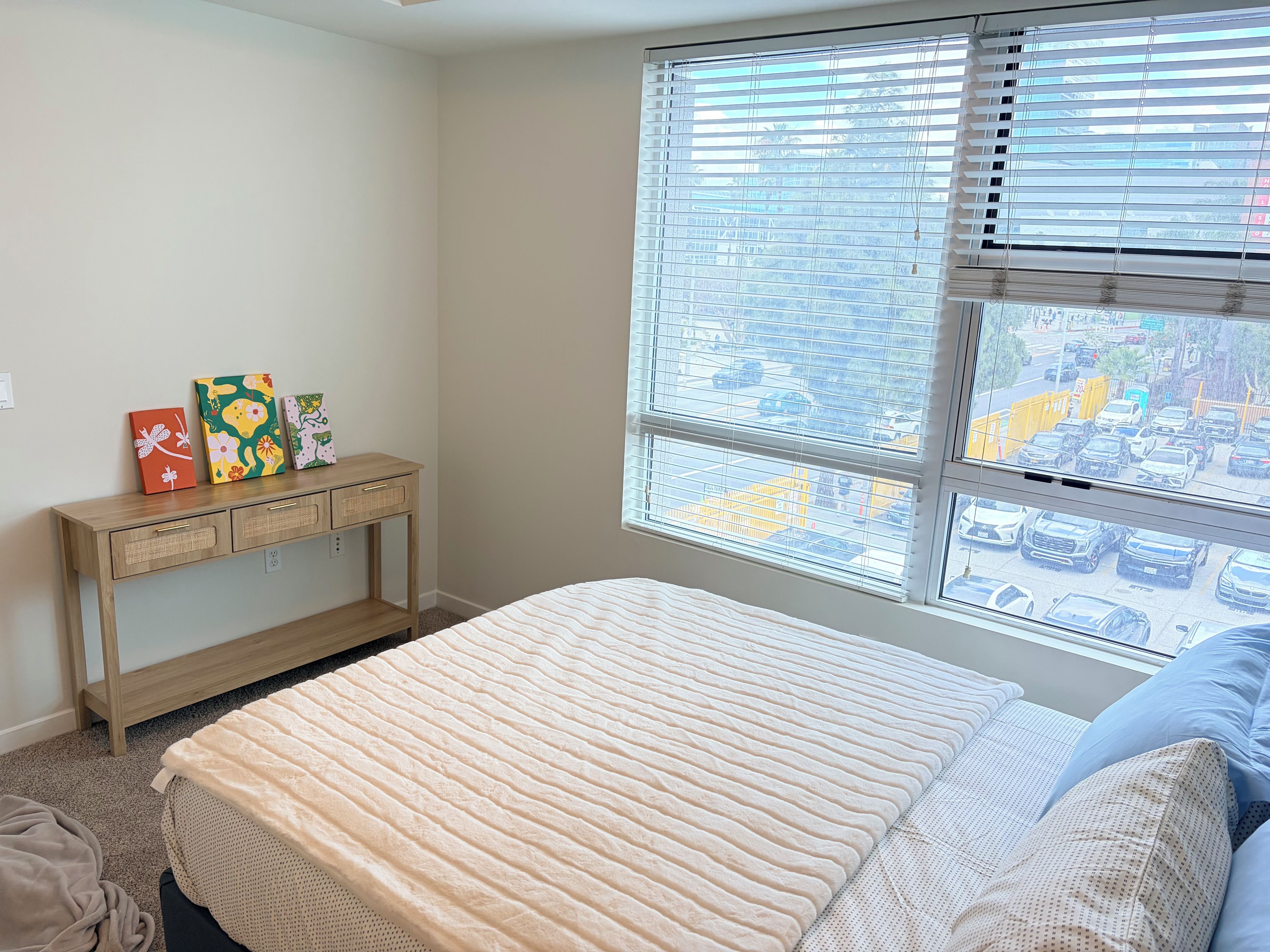 A bedroom with a bed, a bedside table with decorative items, and a window with a view of the street.