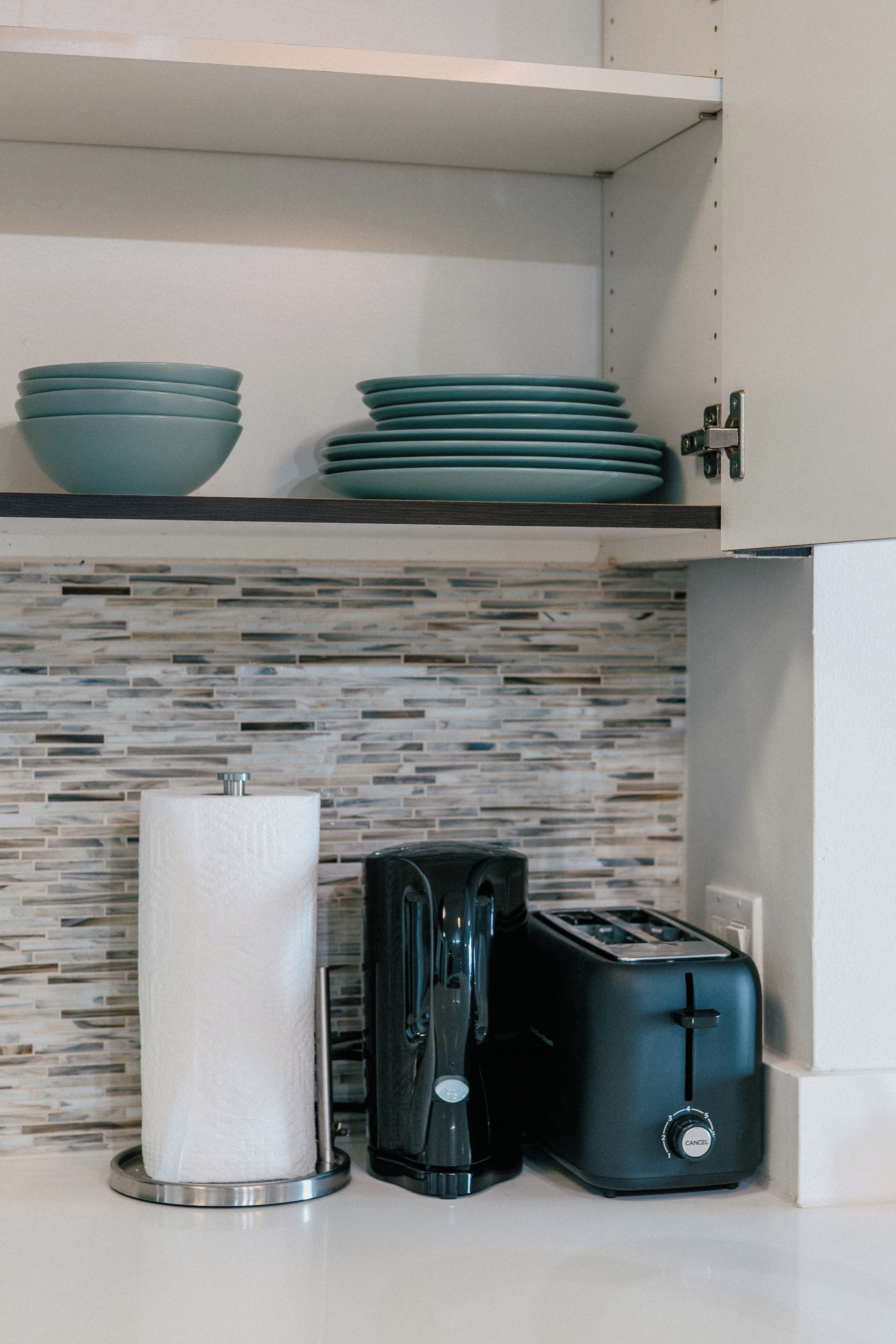 A kitchen with a tiled backsplash, a stack of teal plates, a paper towel holder, a coffee maker, and a toaster.