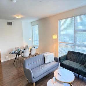 Een moderne woonkamer met een grijze bank, een groene bank, een ronde salontafel en een eethoek met een tafel en stoelen.
