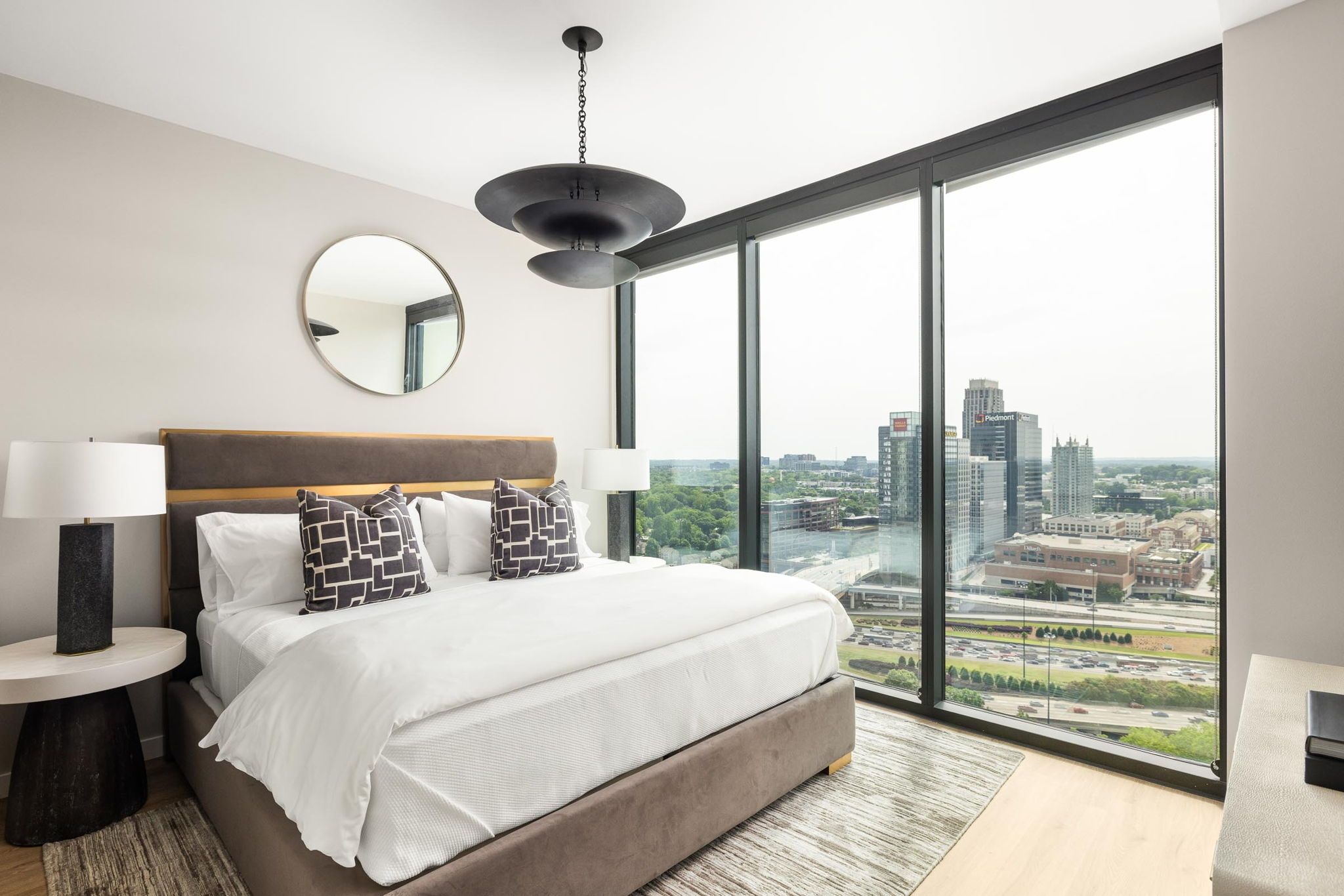 A modern bedroom with a large bed, a round mirror, two bedside lamps, and a city view through floor-to-ceiling windows.