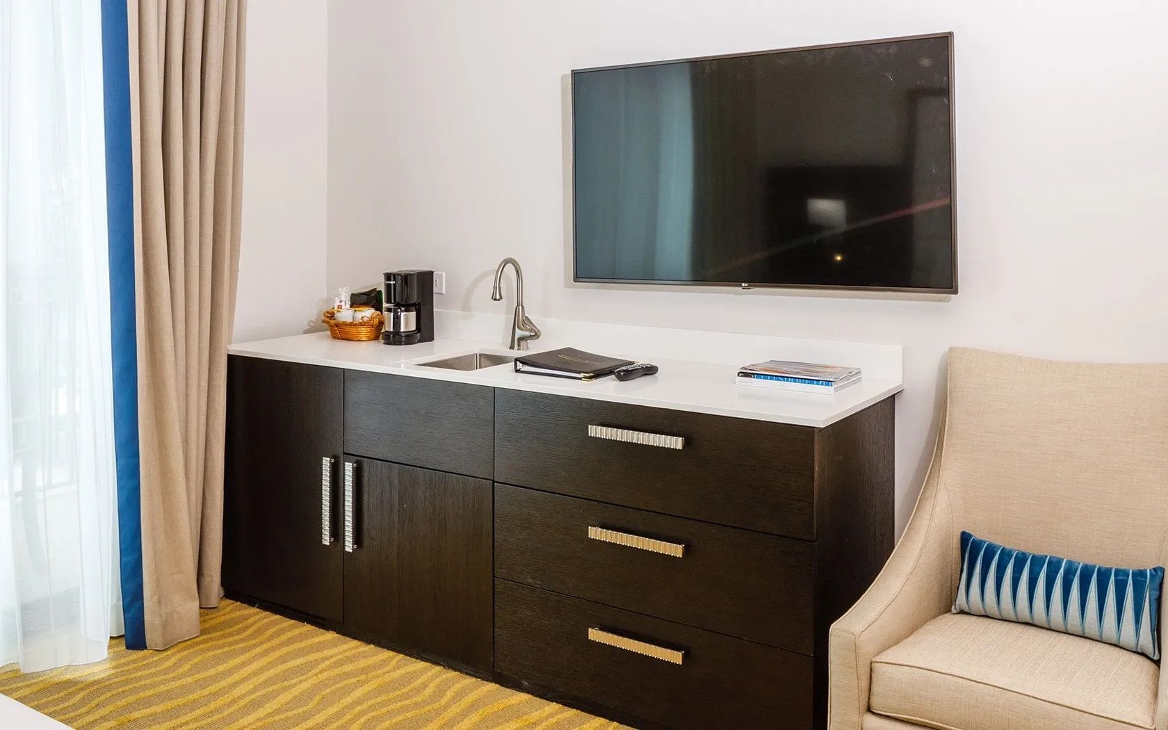 A compact hotel room with a kitchenette, a flat-screen TV, a sofa with a striped pillow, and a window with curtains.