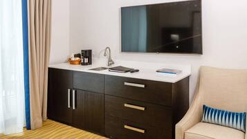 A compact hotel room with a kitchenette, a flat-screen TV, a sofa with a striped pillow, and a window with curtains.