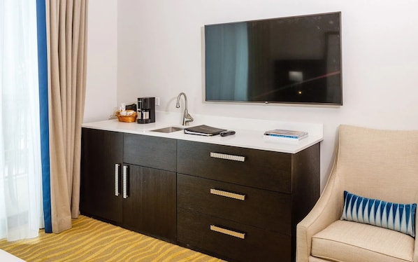 A compact hotel room with a kitchenette, a flat-screen TV, a sofa with a striped pillow, and a window with curtains. - Accessible King Room w/ Marina View | Pools & Spa (Naples)
