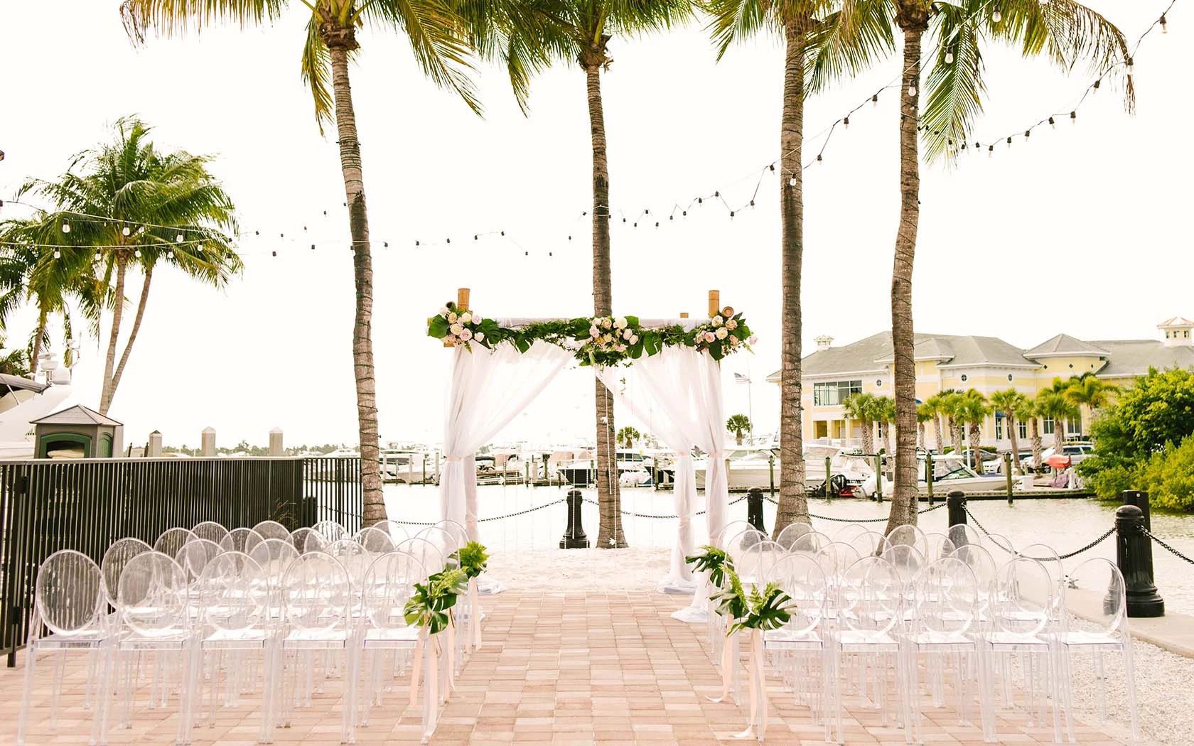 A wedding ceremony setup with a white arch and floral decorations, clear chairs arranged in rows, and palm trees in the background.