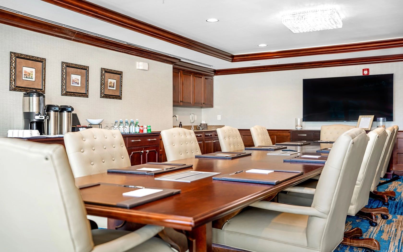 A conference room with a long table, multiple chairs, a coffee station, framed pictures on the wall, and a flat-screen TV.