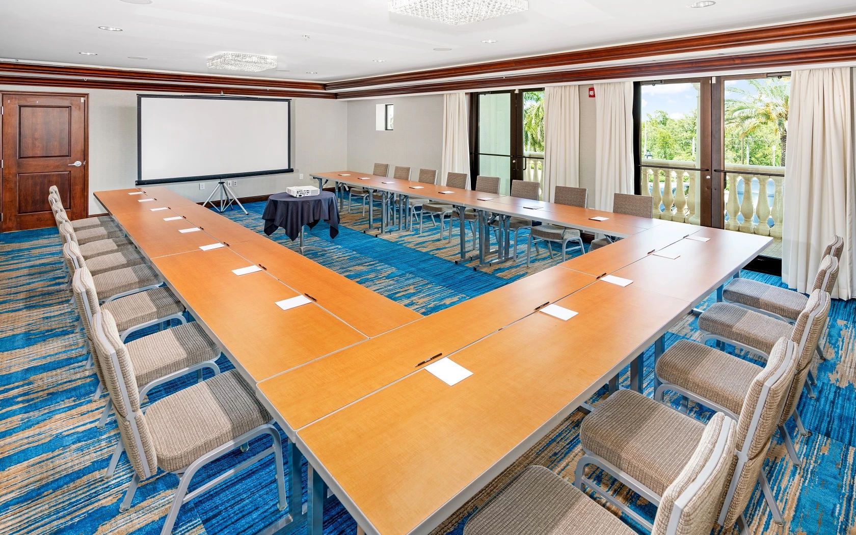 A conference room with a long table, chairs, a projector screen, and a view of the outdoors.