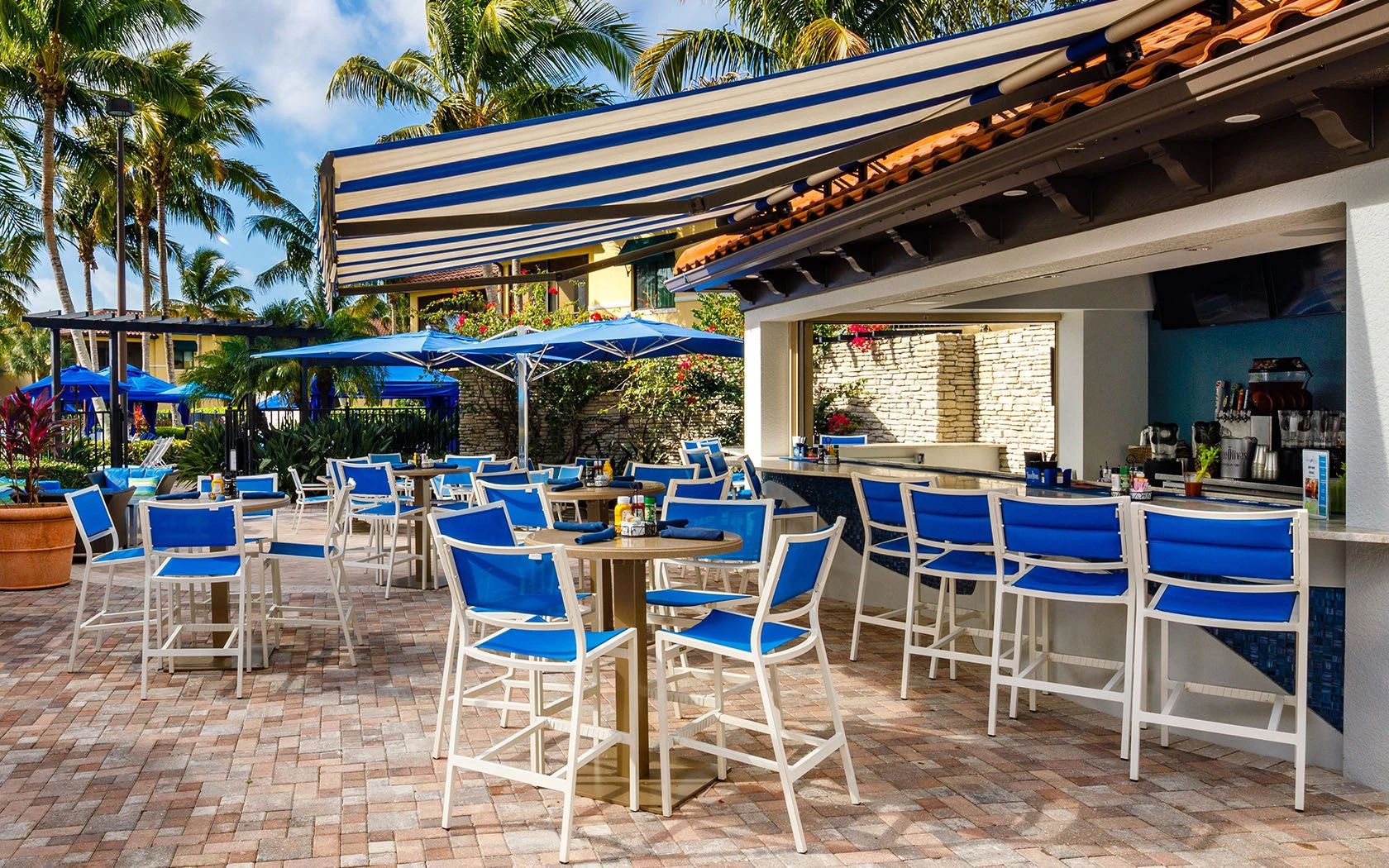 An outdoor dining area with blue chairs and tables, a bar with a stone wall, and palm trees in the background.