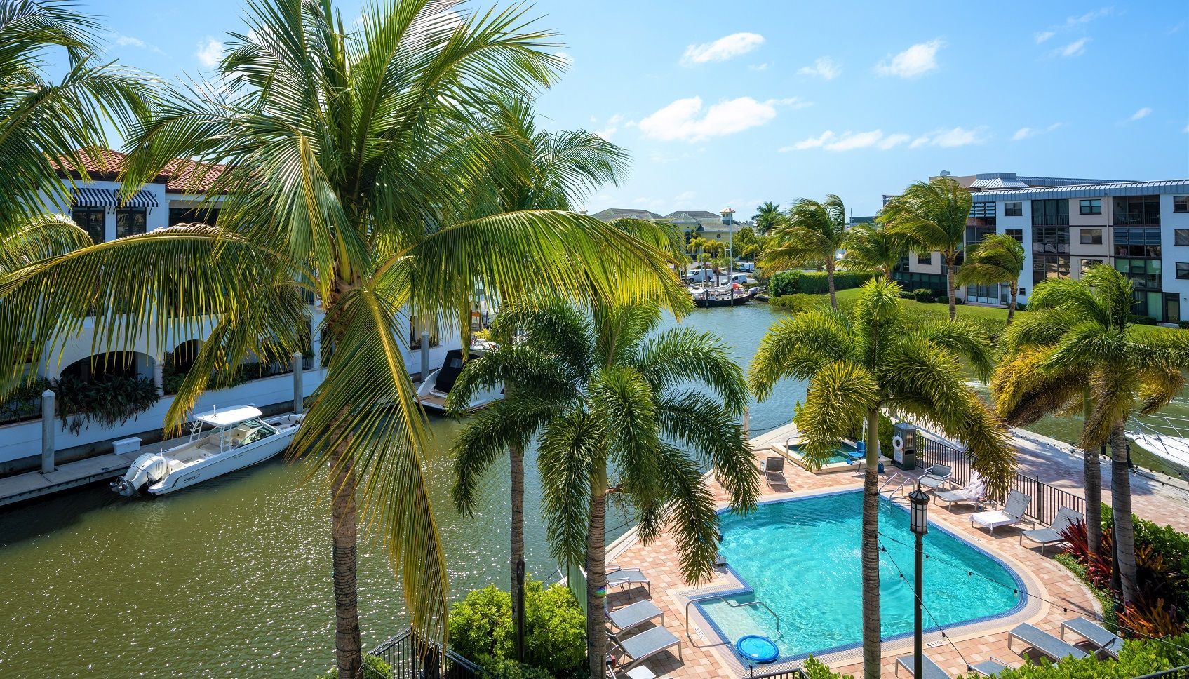 A pool area with palm trees, a boat docked by the water, and a view of a marina with boats.