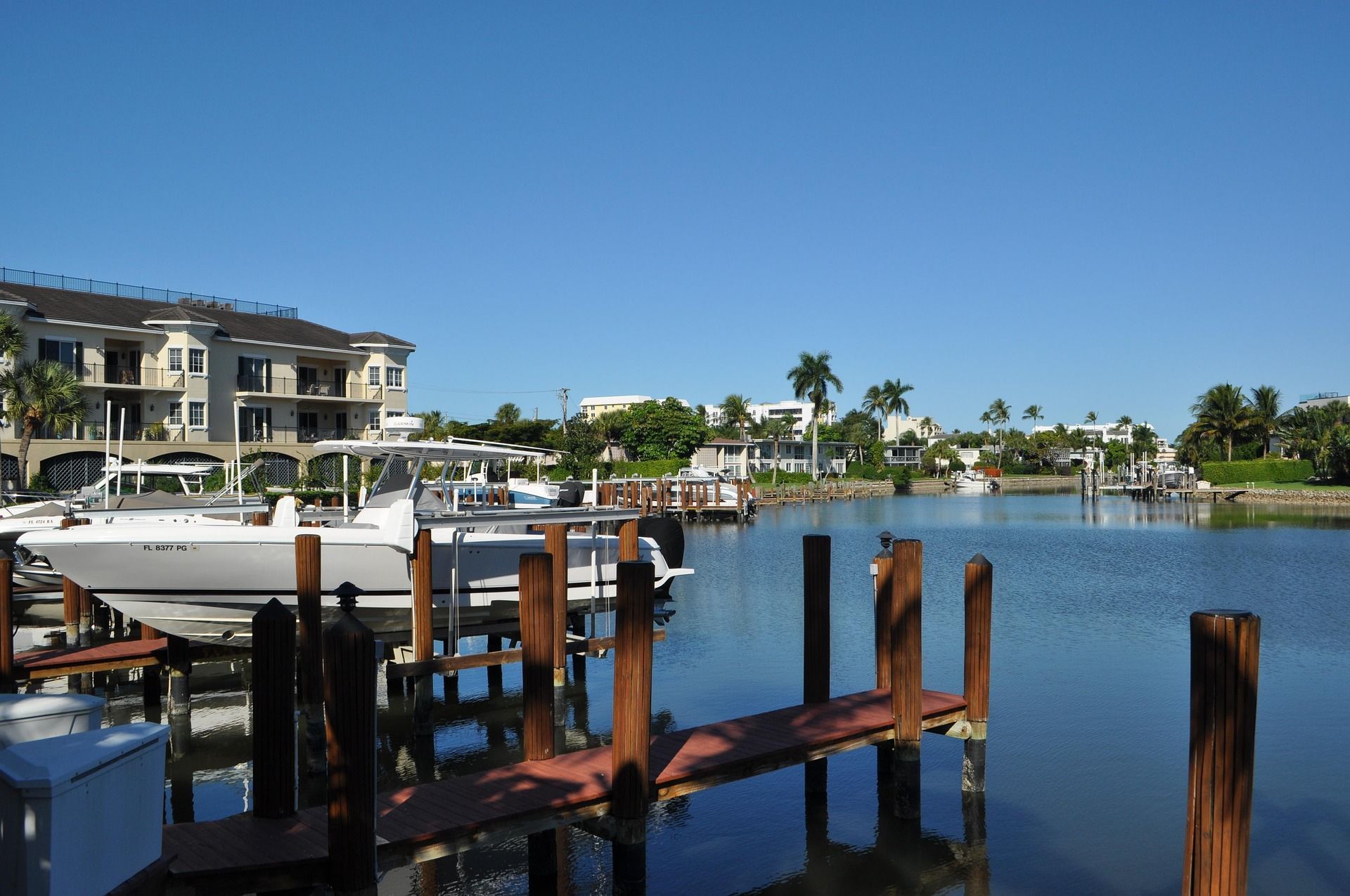 A marina with boats docked, a wooden dock, and a large building with balconies in the background.