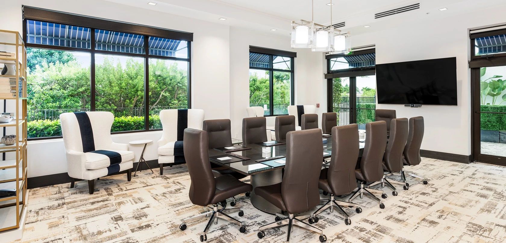 A conference room with a glass table, multiple chairs, a TV, and large windows with a view of greenery.