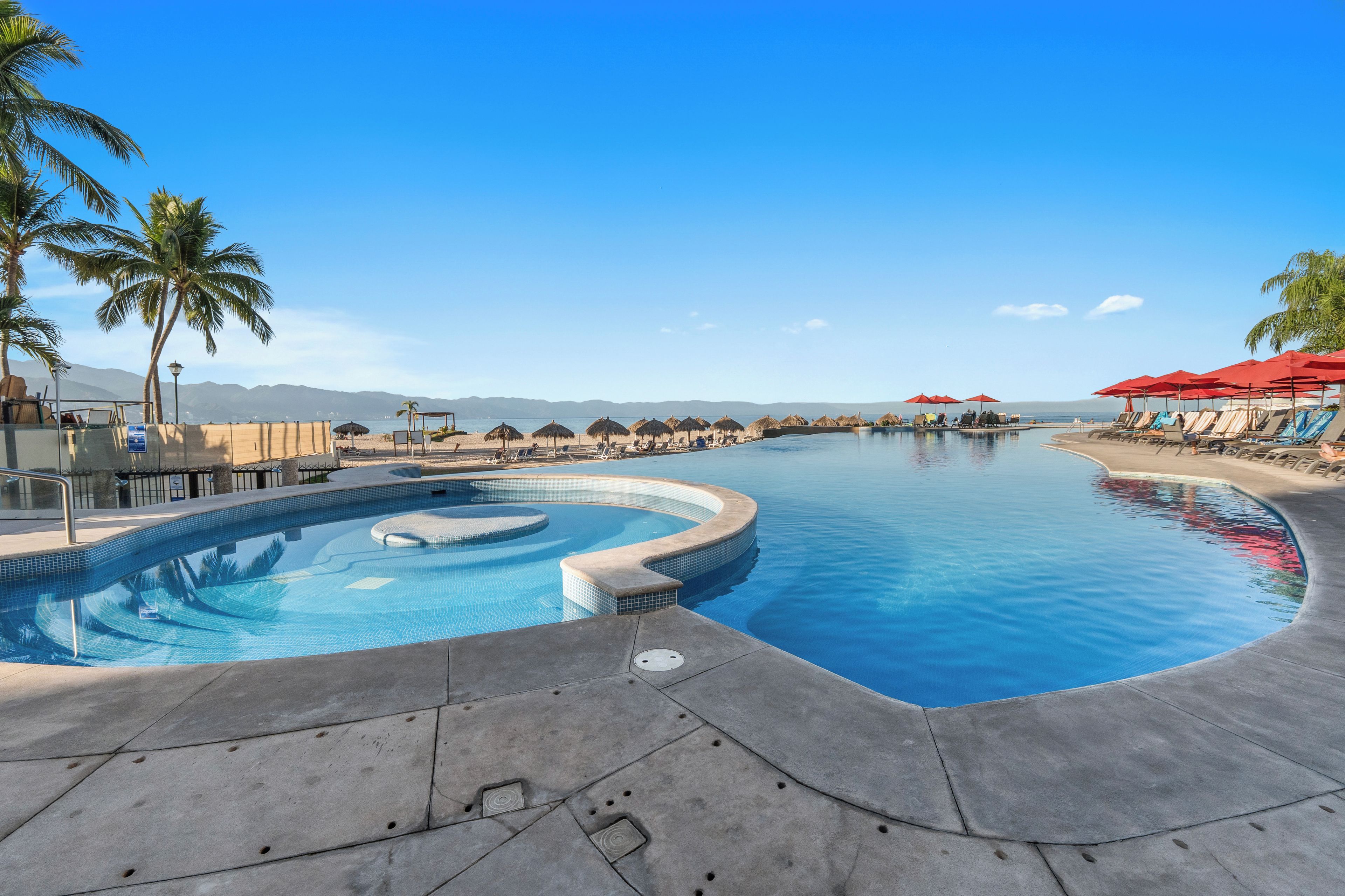 Ein Pool mit gebogener Form, umgeben von Liegestühlen und Sonnenschirmen, mit Palmen und Bergen im Hintergrund.