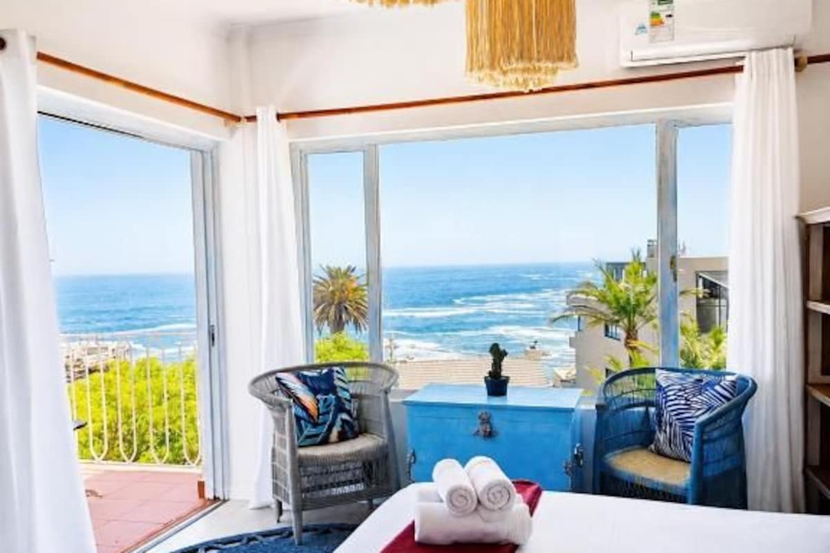 Habitación de hotel con cama, vistas al mar, balcón con una silla y una lámpara colgante.