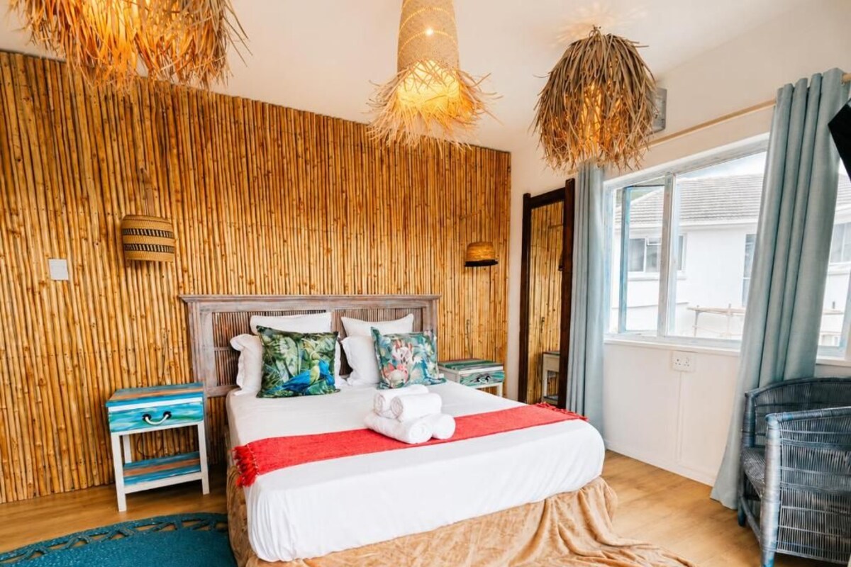 Una habitación de hotel con una cama, mesitas de noche, una silla de mimbre y un ventanal grande.