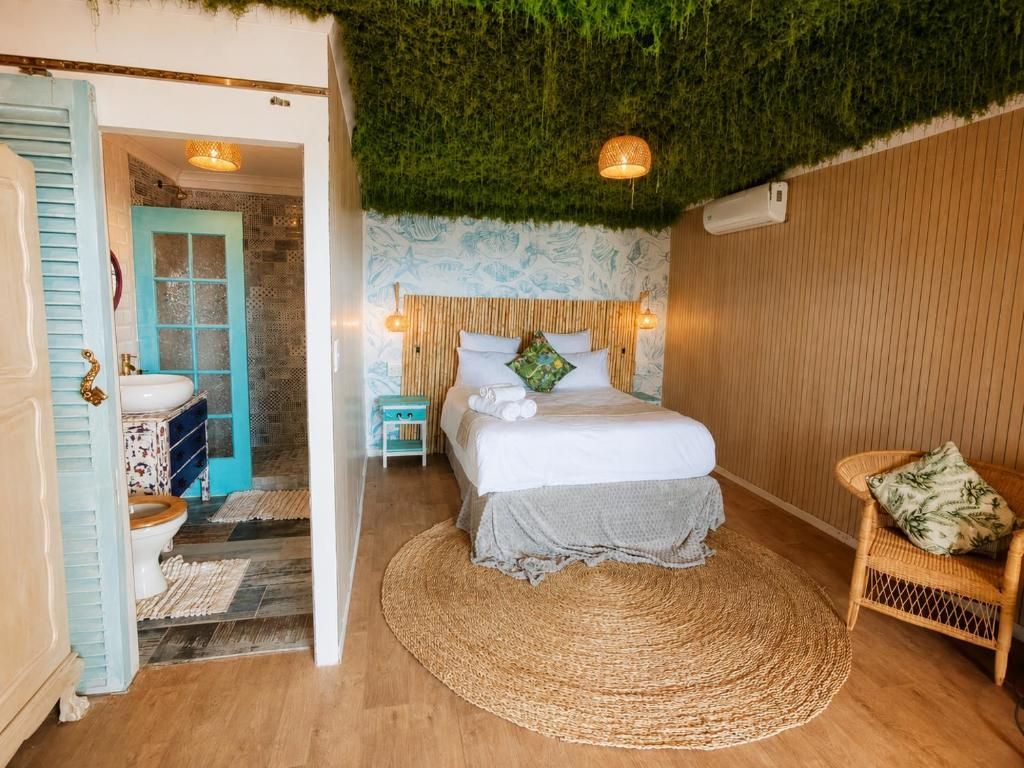 A room with a bed, a wicker chair, a wooden floor, a wall with a textured pattern, and a door leading to another room.