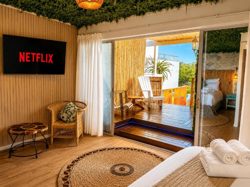 Une pièce avec un téléviseur affichant le logo « NETFLIX », un fauteuil en osier, une petite table ronde, un tapis tissé et un lit avec des serviettes enroulées.