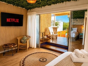Habitación con una televisión mostrando 'NETFLIX', una silla de mimbre, una mesita redonda pequeña, un tapete tejido y una cama con toallas dobladas.