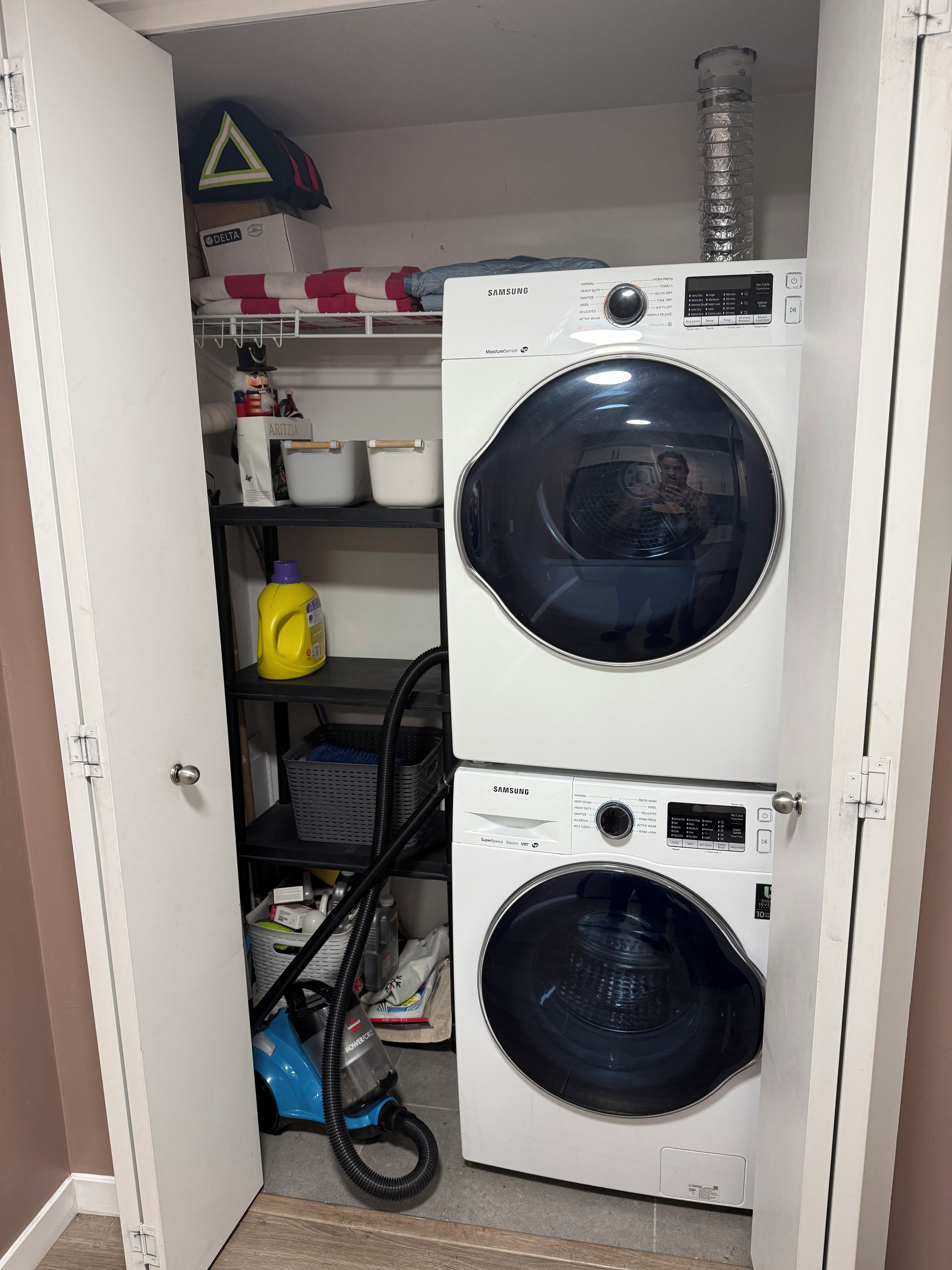 A laundry room with two stacked front-loading washing machines, a shelf with various items, and a vacuum cleaner.