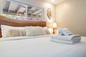 Une chambre d’hôtel avec un lit, un attrape-rêves accroché au mur, une fenêtre donnant sur des arbres et une lampe.