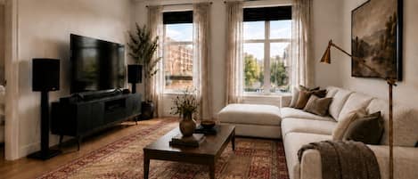 A living room with a sofa, a coffee table, a TV, and a large window with curtains.