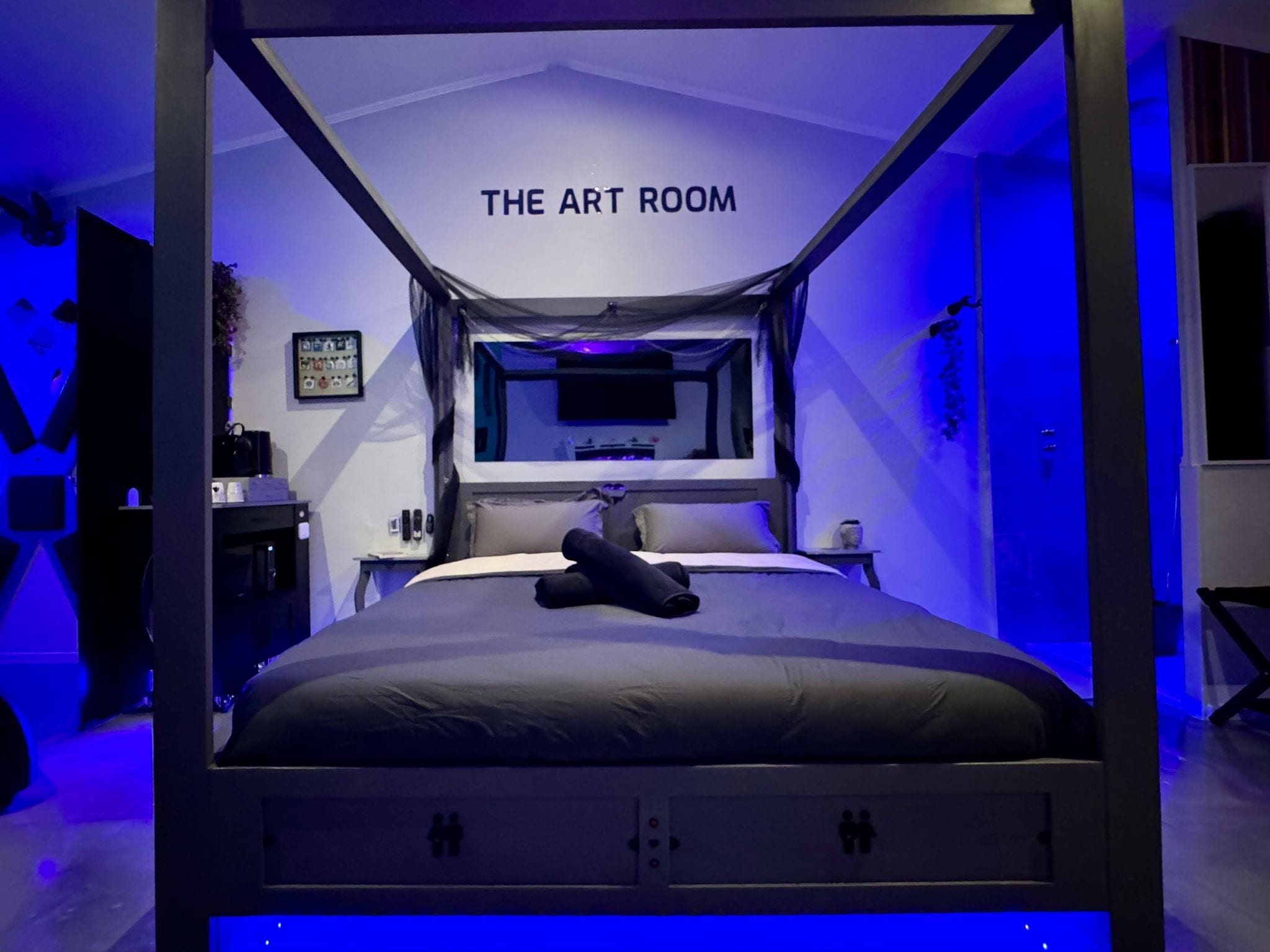 Una cama con dosel y cortinero, con un letrero que dice 'THE ART ROOM' en un dormitorio moderno con televisor de pantalla plana y una mesita.