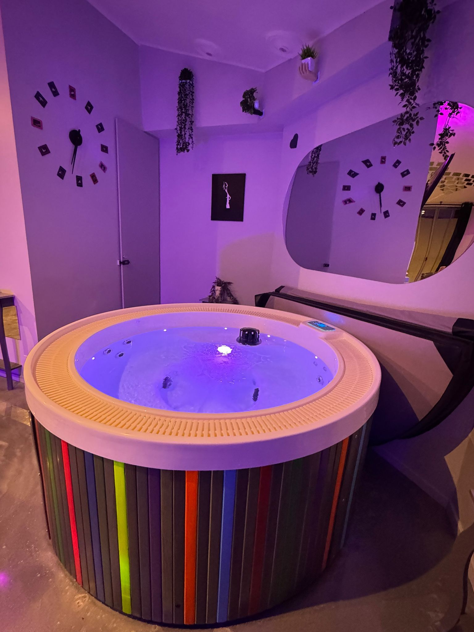 Una habitación con un jacuzzi grande y circular en el centro, rodeado por un muro a rayas. Hay relojes en las paredes y un espejo sobre el jacuzzi.