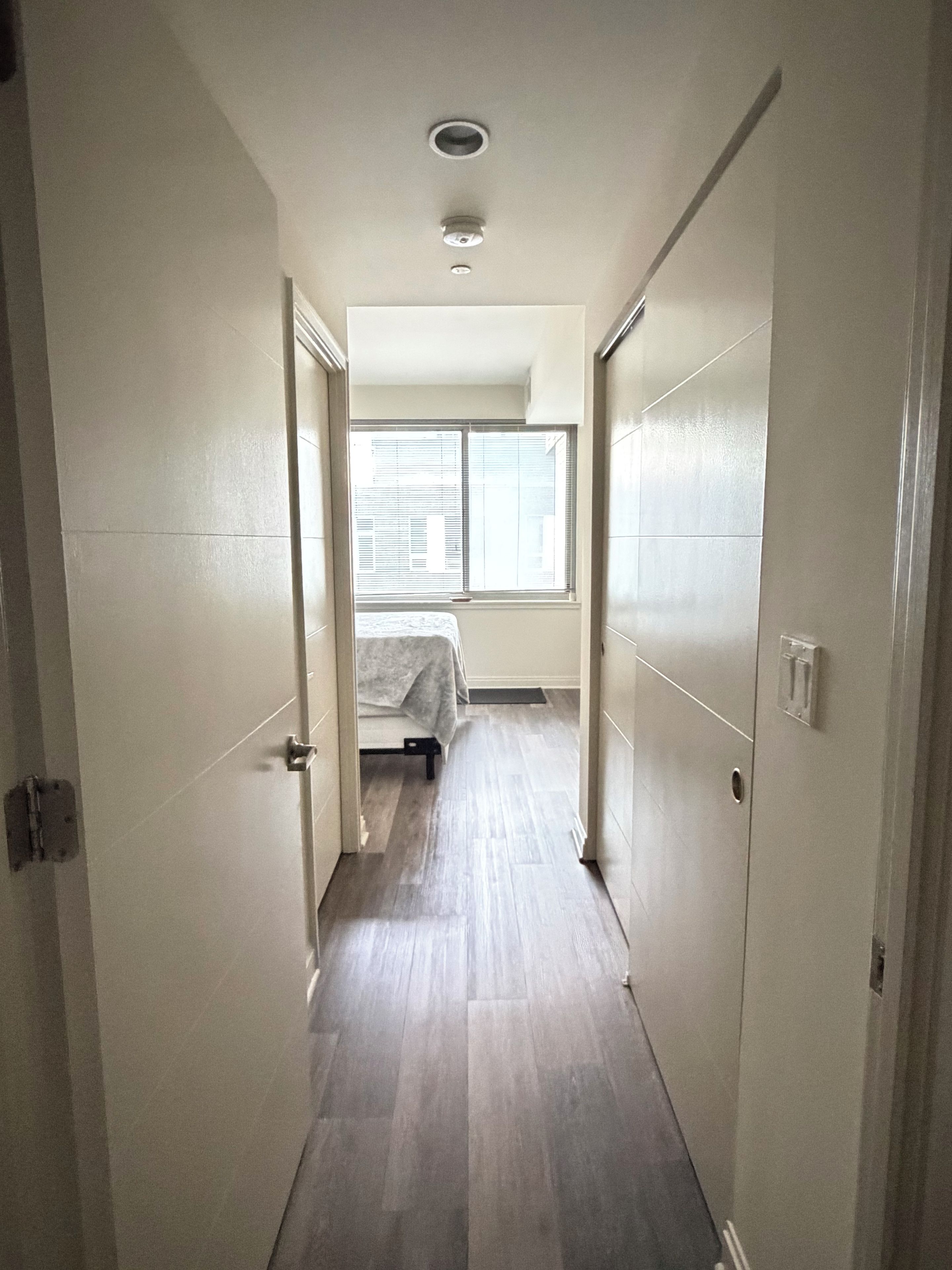 Un pasillo estrecho con una cama al fondo, una ventana con persianas y una puerta a la izquierda.