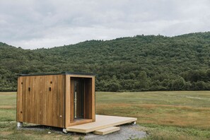Eine Holzhütte mit Tür und einer kleinen Holzplattform in einem grasbewachsen Feld, im Hintergrund ein bewaldeter Hügel.
