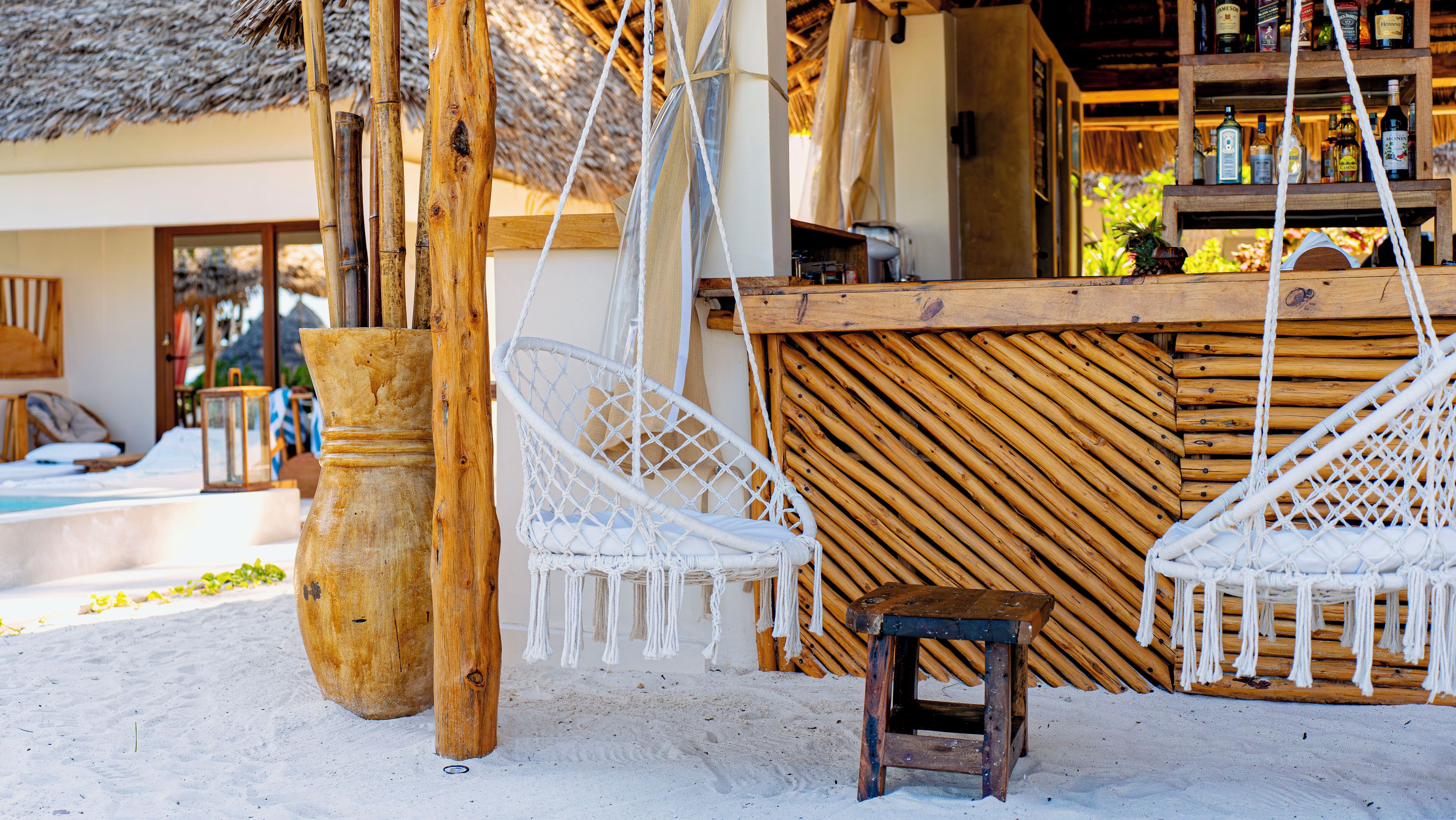A beachside bar area with a wooden counter, two white wicker chairs, and a small wooden stool.