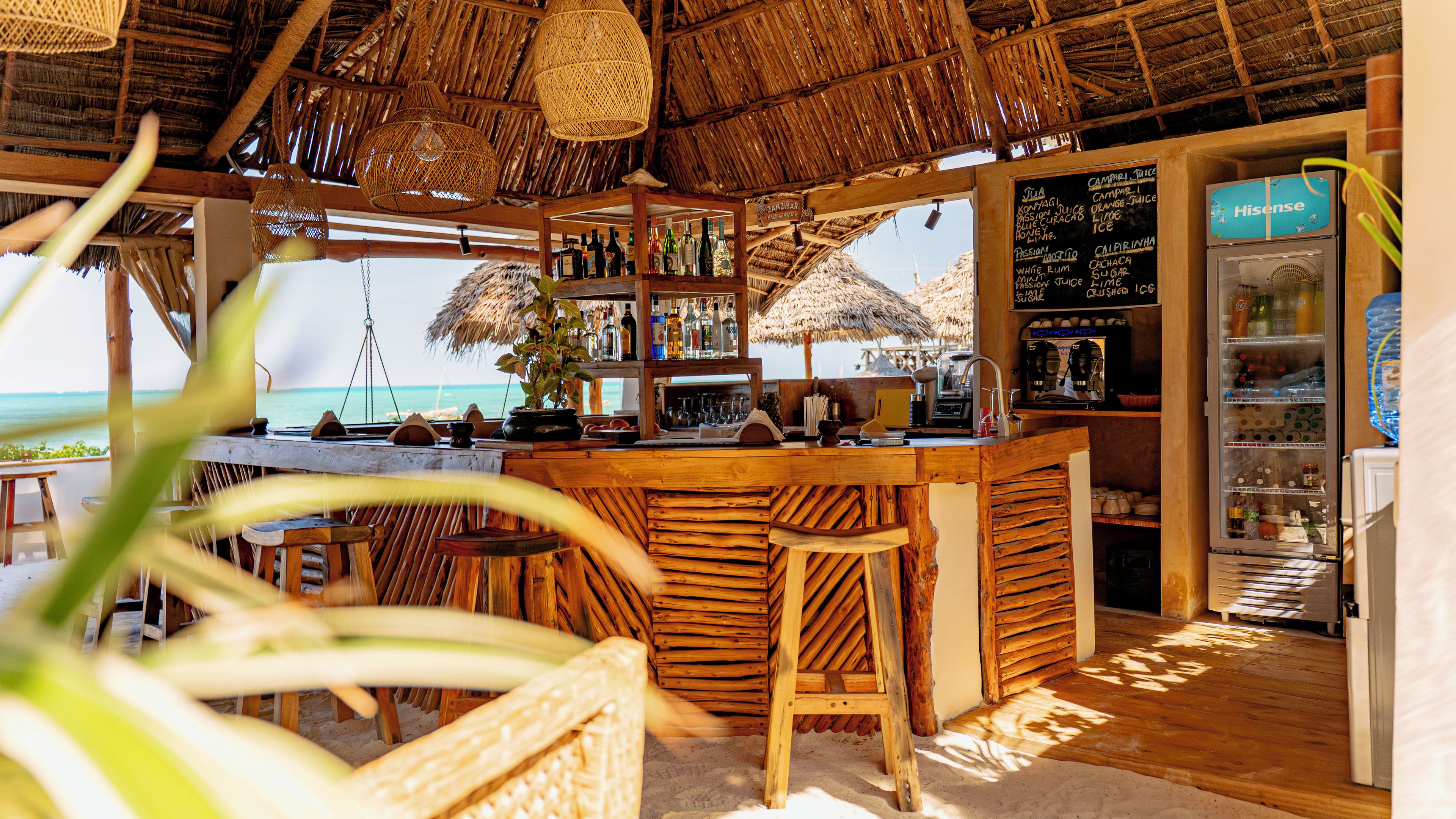 Un bar en la playa con mostrador de madera, taburetes y vista al océano.