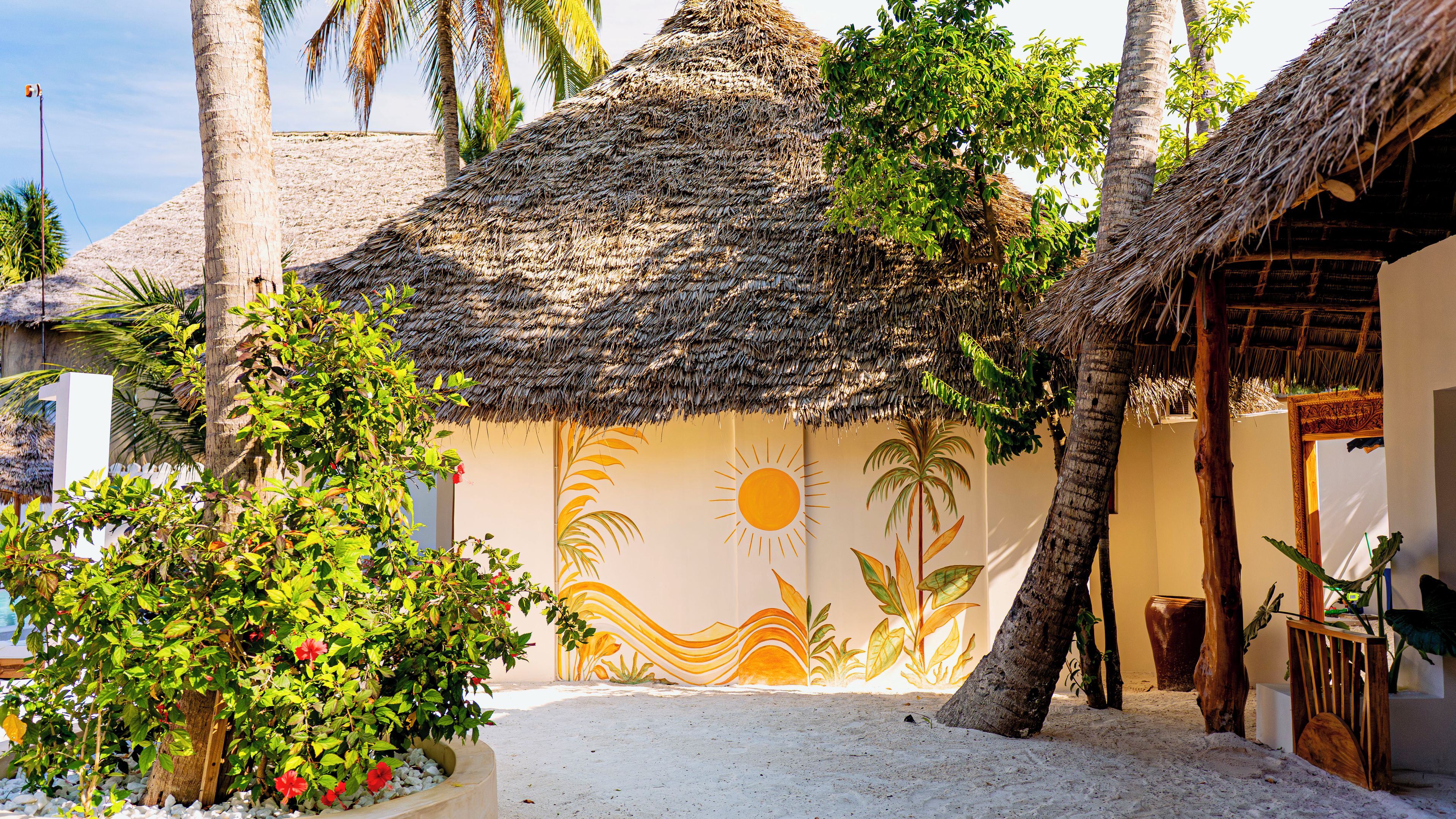 A beachside building with a thatched roof, surrounded by palm trees and a sandy ground.