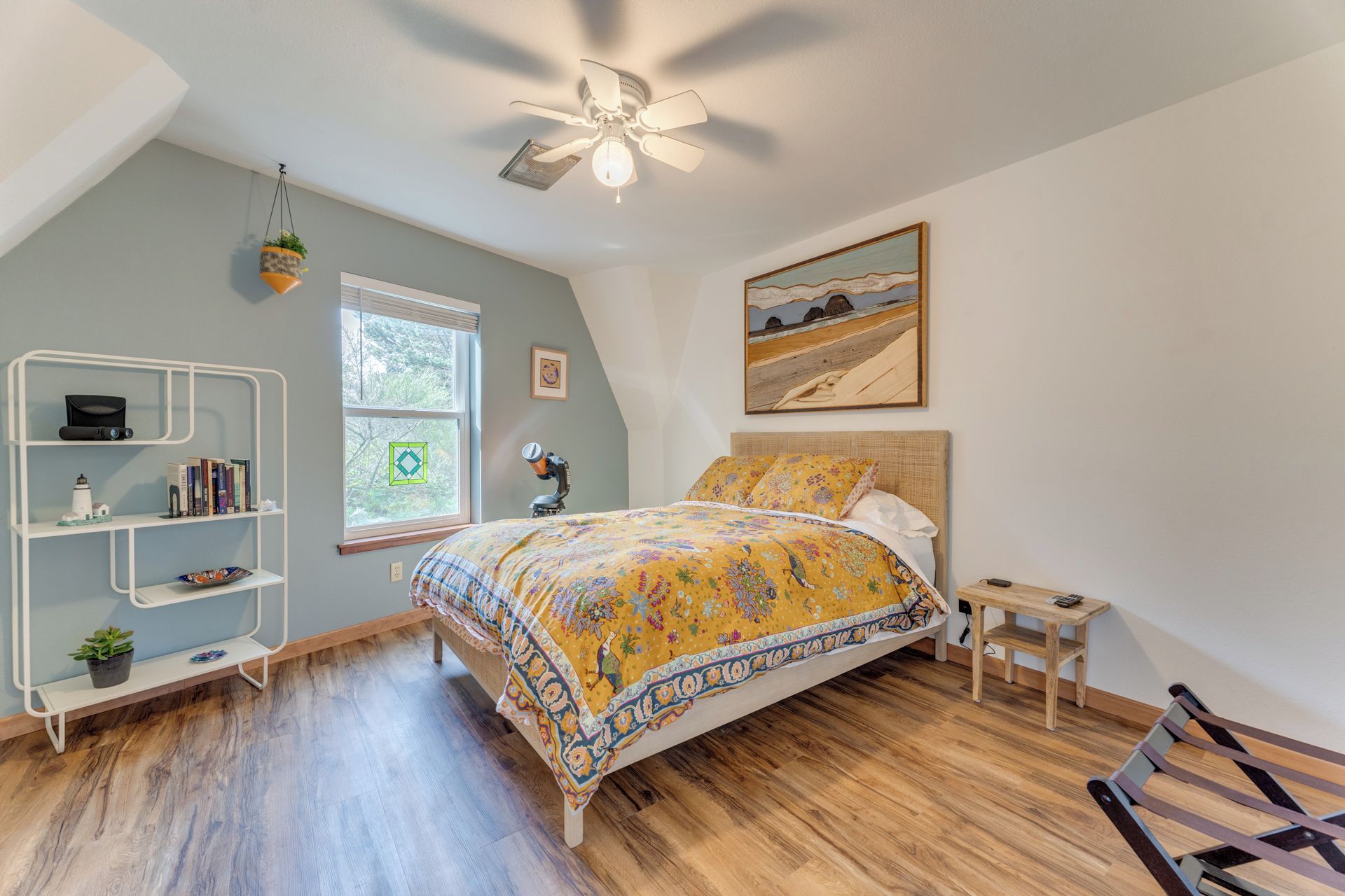 A bedroom with a bed, a desk, a bookshelf, a ceiling fan, and a window.