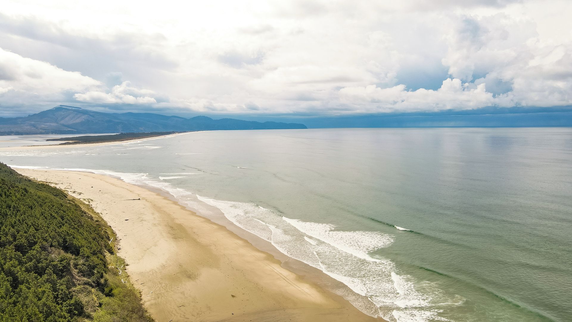 A coastal landscape with a sandy beach, calm sea, and distant mountains.