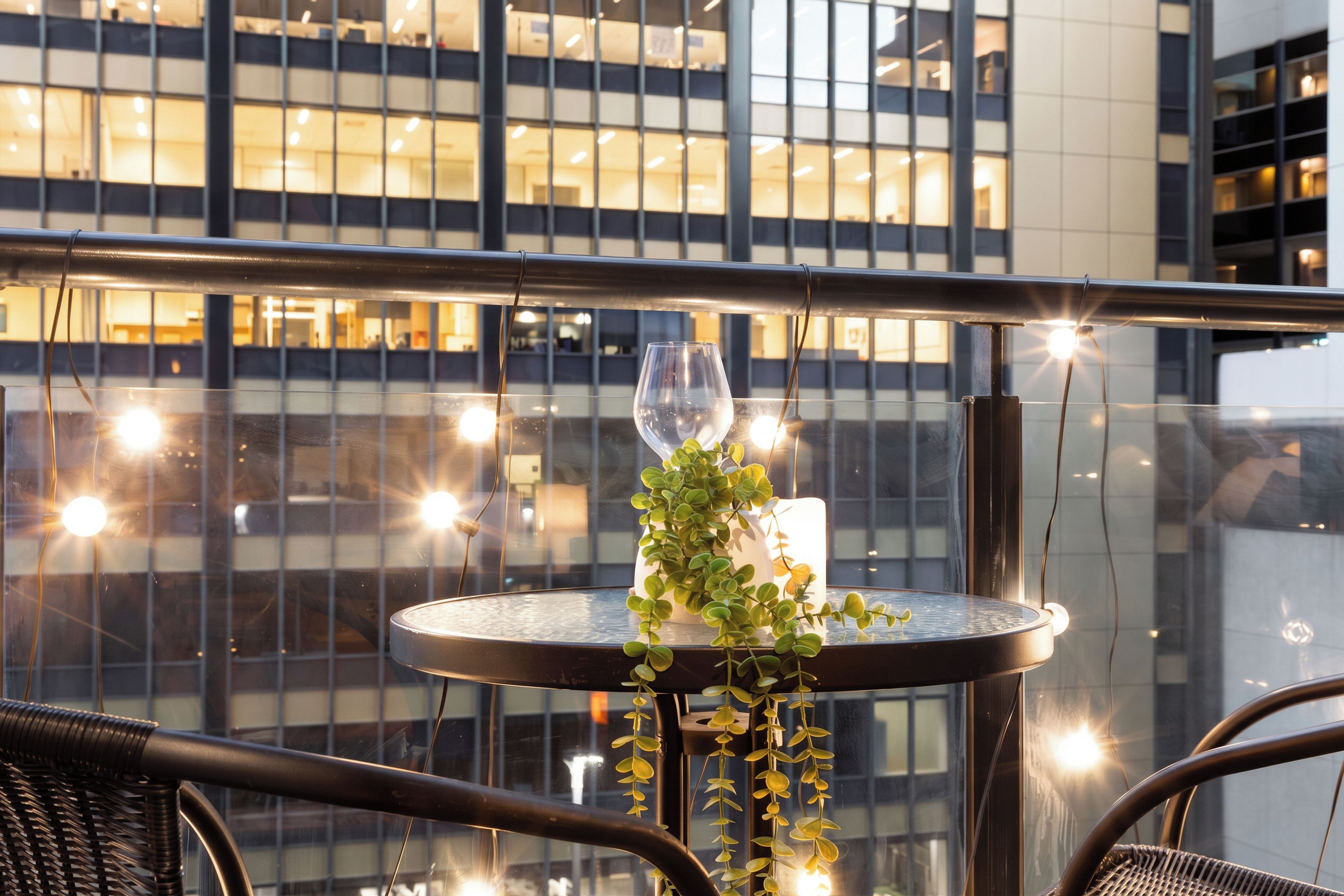 Een ronde tafel met een glas, een kaarsje en een plant op een balkon met uitzicht op een gebouw met veel ramen.