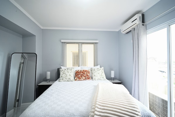 Un dormitorio con una cama, dos mesitas de noche, un ventanal con persianas, un equipo de aire acondicionado y vistas al exterior.