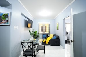Un espacio habitable compacto con comedor, un sofá con cojines, un televisor montado en la pared y una ventana con persianas.