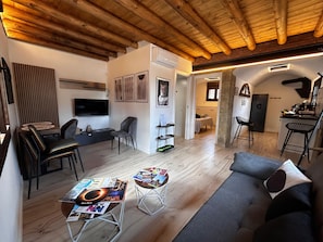 Een moderne woonruimte met een houten plafond, een flatscreen televisie, een grijze bank en een eethoek met een tafel en stoelen.