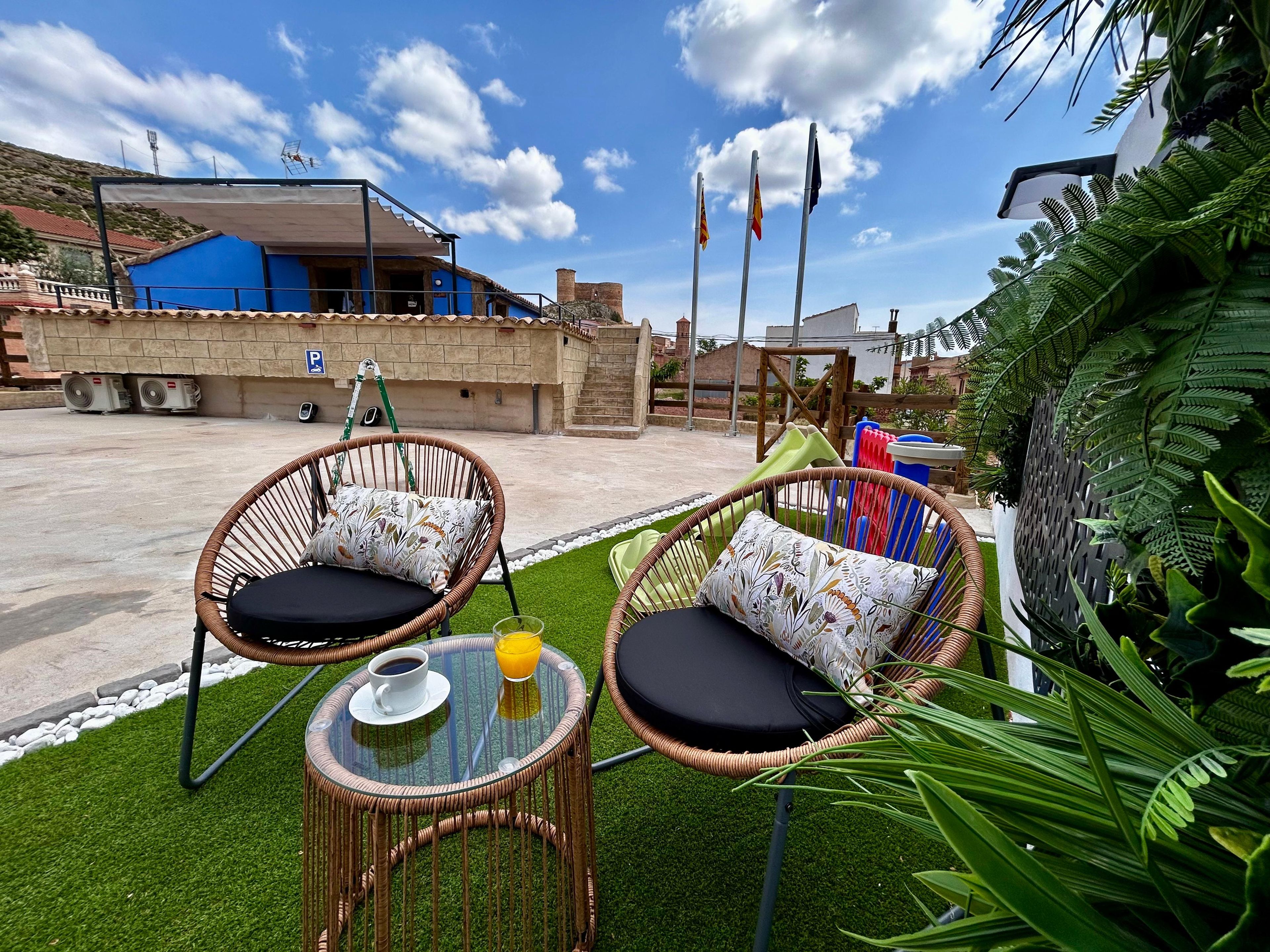 An outdoor seating area with wicker chairs and a table, surrounded by greenery and a clear sky.
