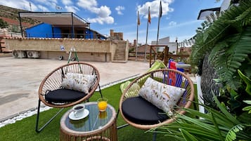 An outdoor seating area with wicker chairs and a table, surrounded by greenery and a clear sky.