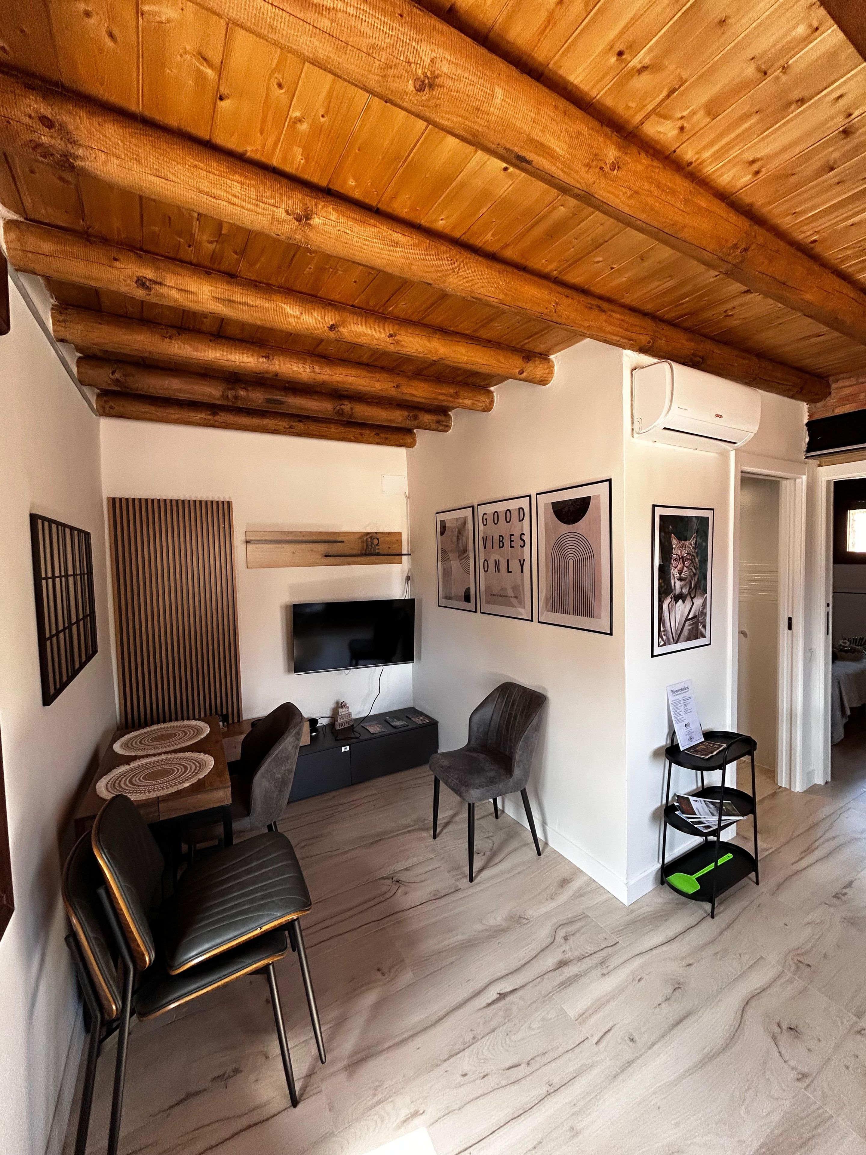 A modern living room with a wooden ceiling, a flat-screen TV, a dining area with a table and chairs, and a small bookshelf.