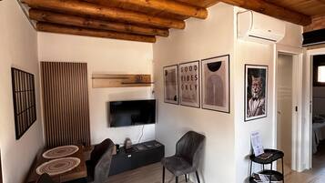 A modern living room with a wooden ceiling, a flat-screen TV, a dining area with a table and chairs, and a small bookshelf.